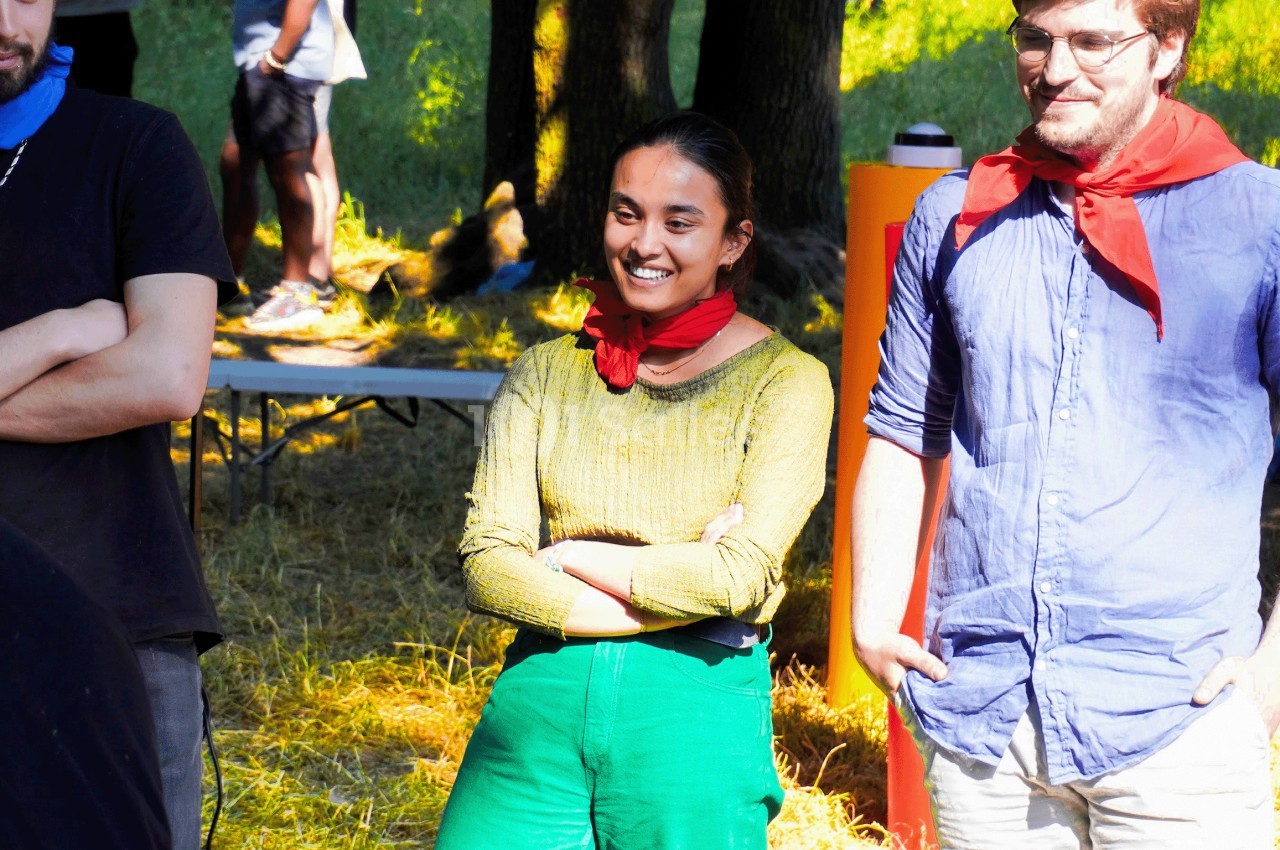 Trois personnes discutent en plein air, souriantes, portant des foulards rouges dans un cadre ensoleillé et boisé.
