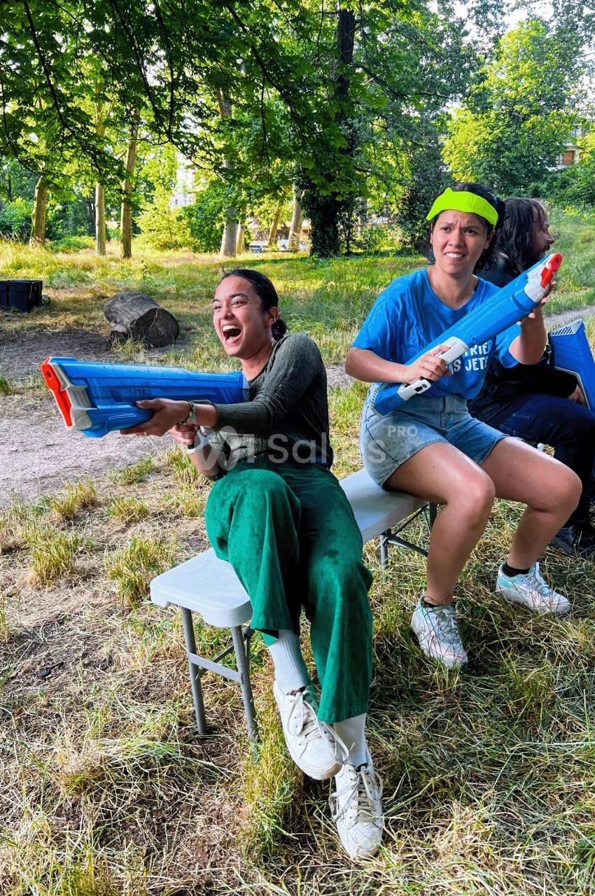 Deux personnes assises sur un banc s'amusent avec des pistolets à eau dans un parc verdoyant.