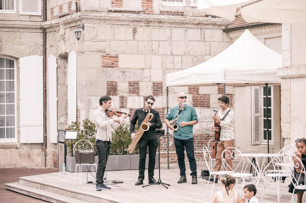 Un groupe de musiciens joue en plein air devant un bâtiment ancien, avec un public assis à proximité.