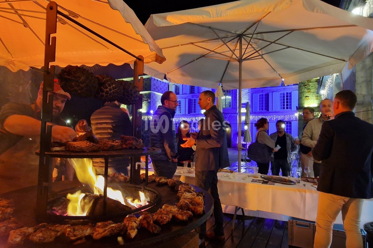 Un chef cuisine des viandes sur un barbecue circulaire en plein air lors d'un événement nocturne sous des parasols éclairés.