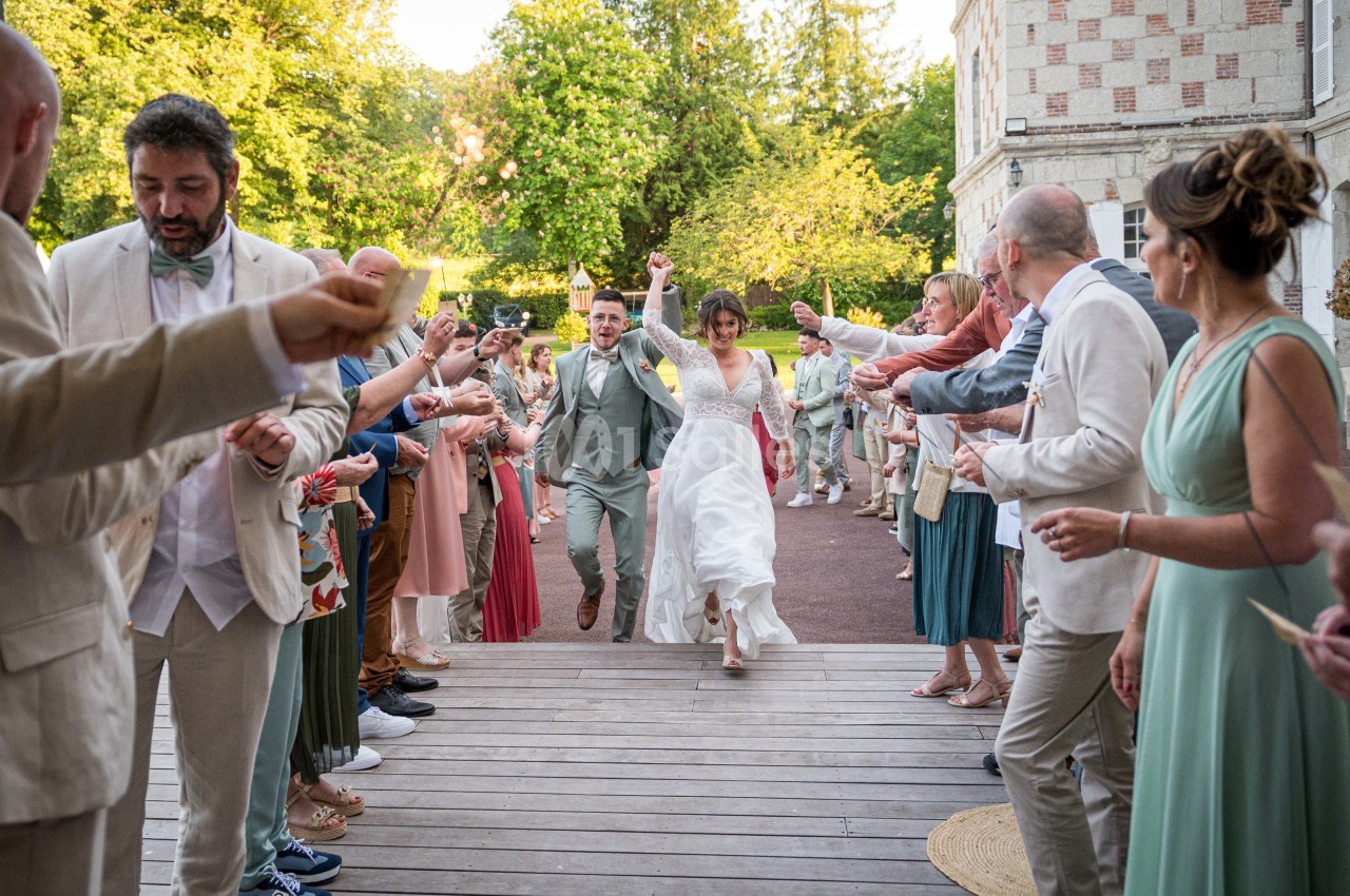 Un couple de mariés traverse une haie d'invités applaudissant sur une terrasse en bois, entourée de verdure.