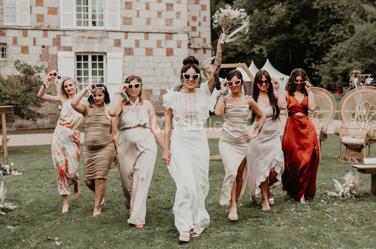 Un groupe de femmes en robes élégantes marche sur une pelouse devant un bâtiment en briques, portant des lunettes fantaisie.