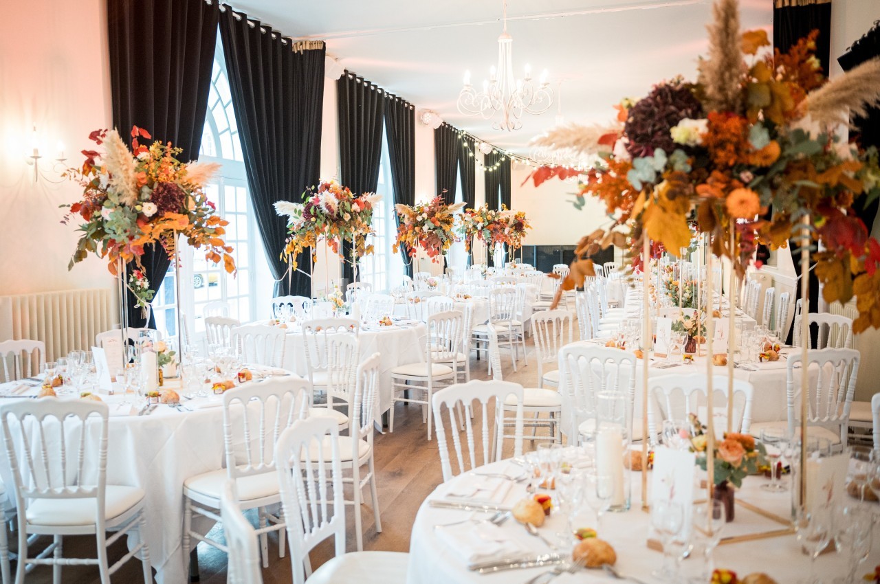 Salle de réception décorée pour un mariage avec des tables rondes, nappes blanches et centres de table floraux aux tons…