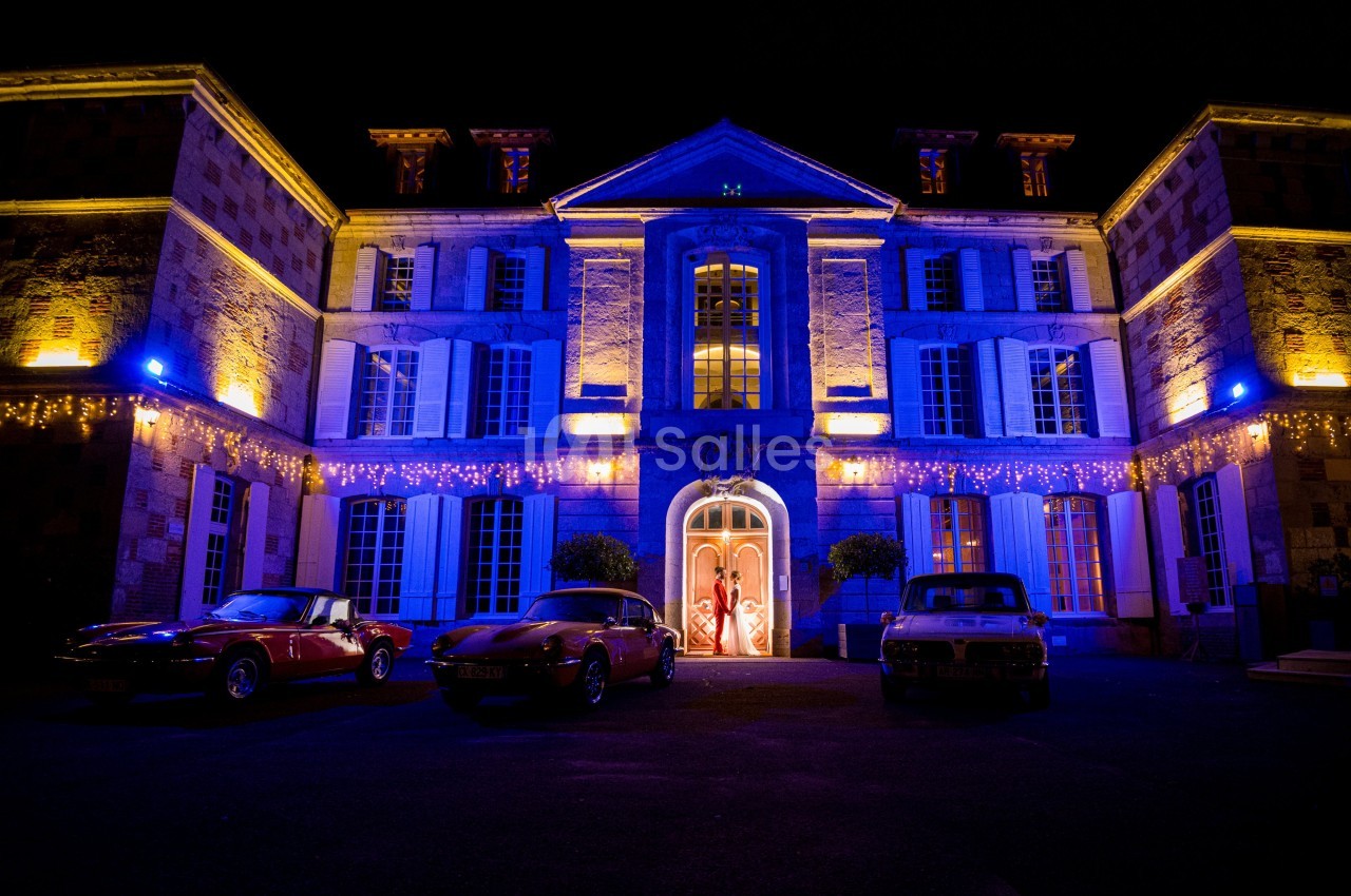Façade d'un bâtiment illuminé en bleu et jaune la nuit, avec trois voitures anciennes stationnées devant.