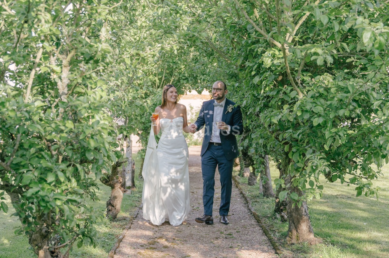 Un couple en tenue de mariage marche sur un sentier bordé d'arbres dans un jardin verdoyant.