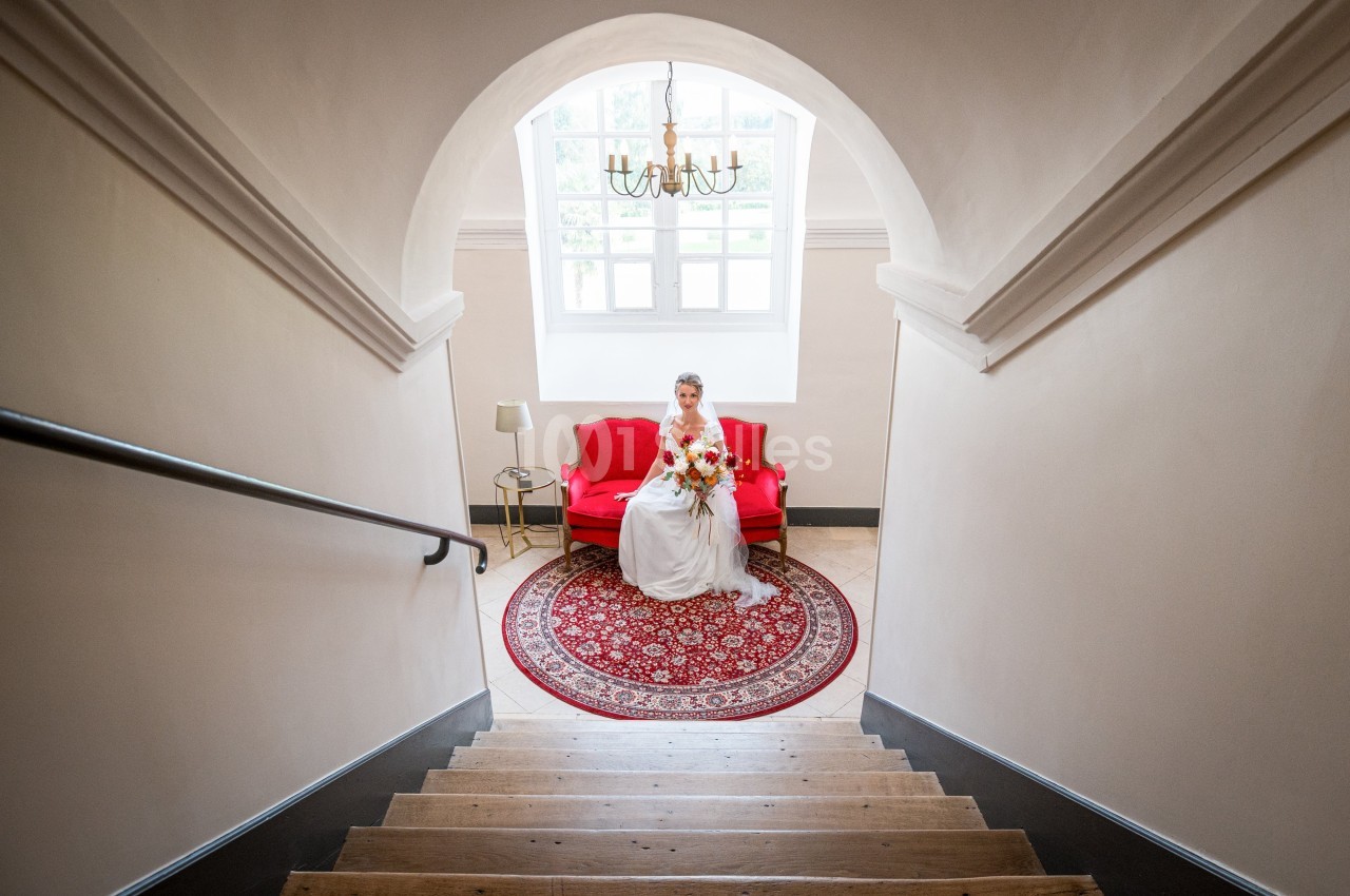 Une mariée en robe blanche est assise sur un canapé rouge, dans une pièce lumineuse avec un tapis rond.