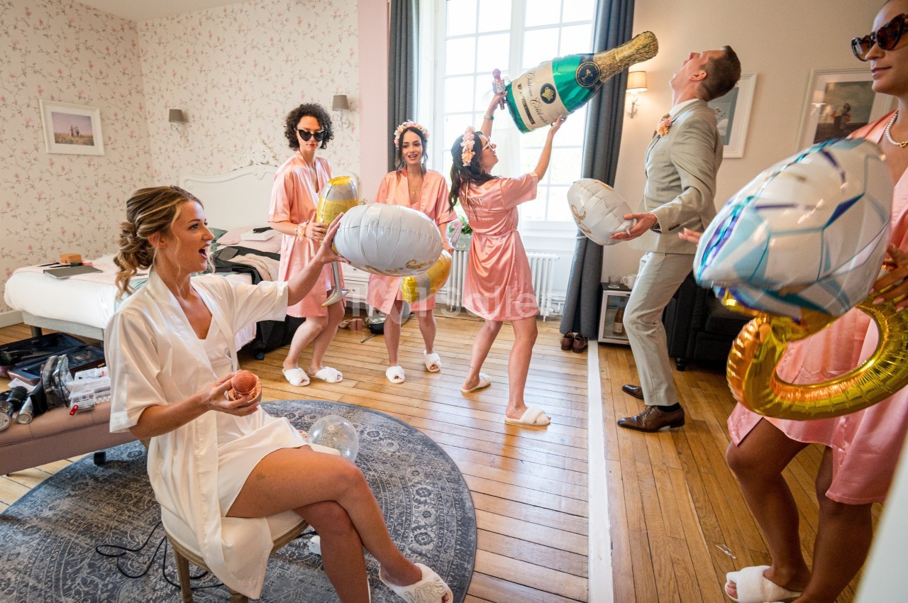 Des amies en peignoirs et un homme en costume s'amusent avec des accessoires gonflables dans une chambre lumineuse.