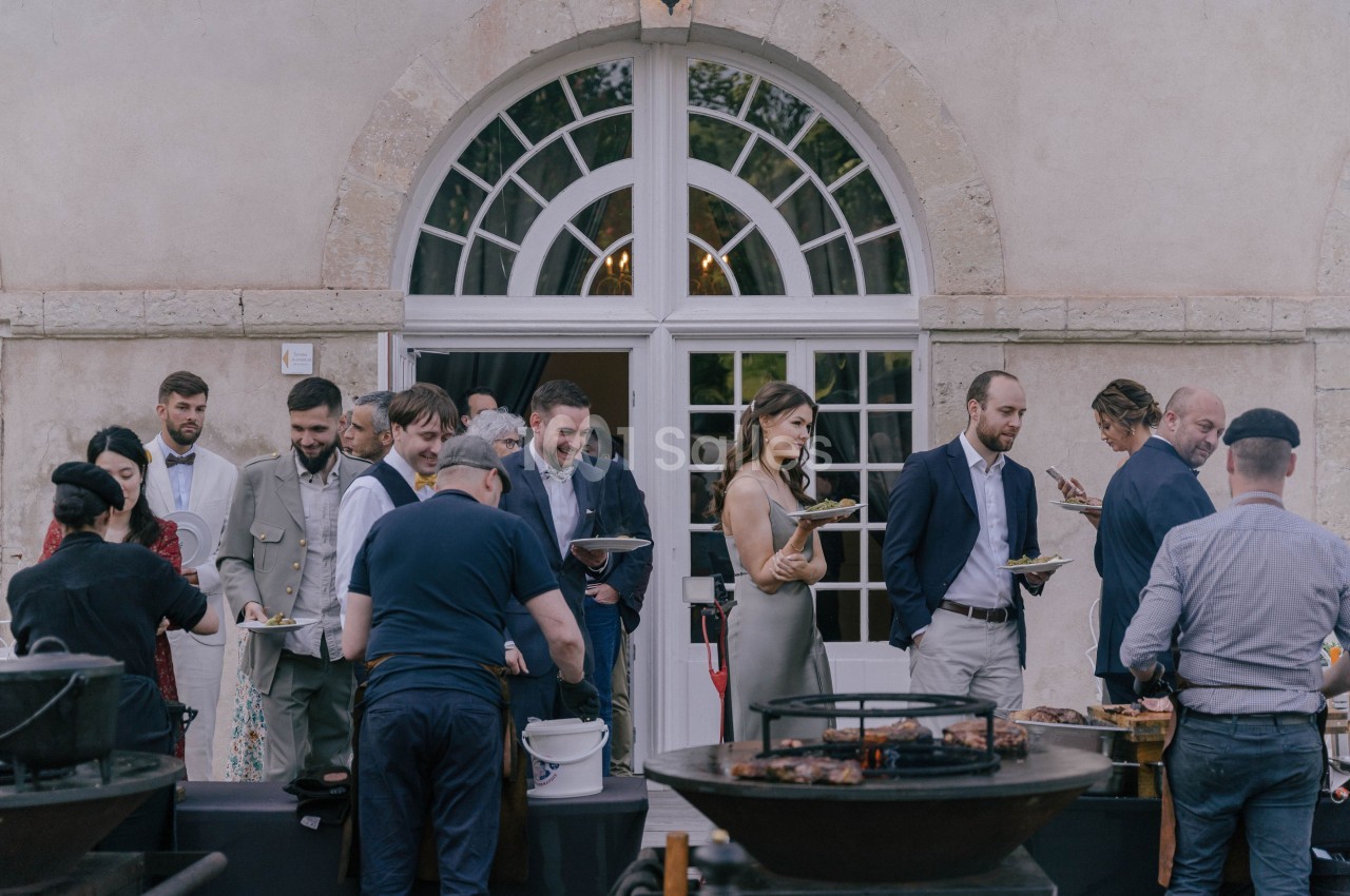 Des personnes rassemblées devant un bâtiment avec des assiettes, près de barbecues en extérieur.