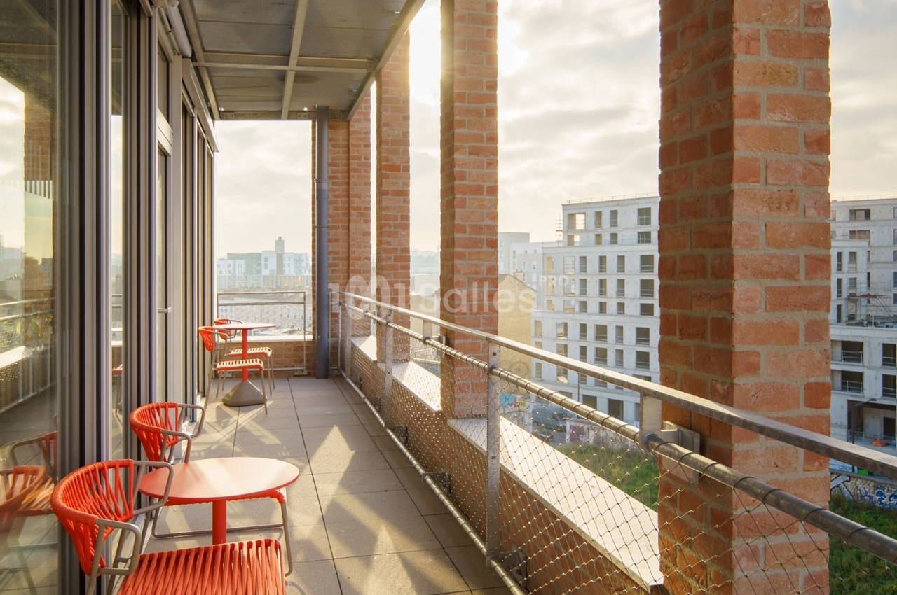 Balcon avec mobilier rouge, encadré de colonnes en briques, offrant une vue sur des immeubles modernes au coucher du soleil.