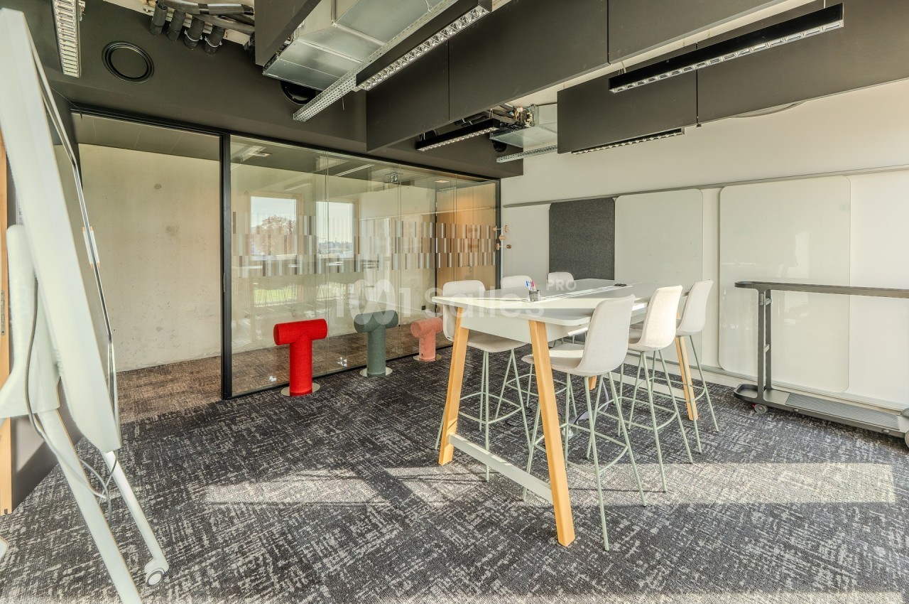 Salle de réunion moderne avec table haute, chaises, tableau blanc et cloison vitrée laissant passer la lumière naturelle.