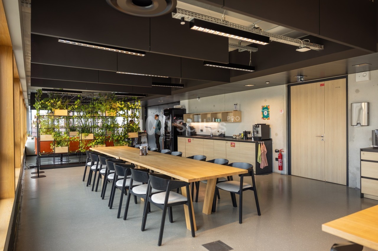 Salle de pause moderne avec tables en bois, chaises noires, kitchenette, plantes suspendues et éclairage naturel.