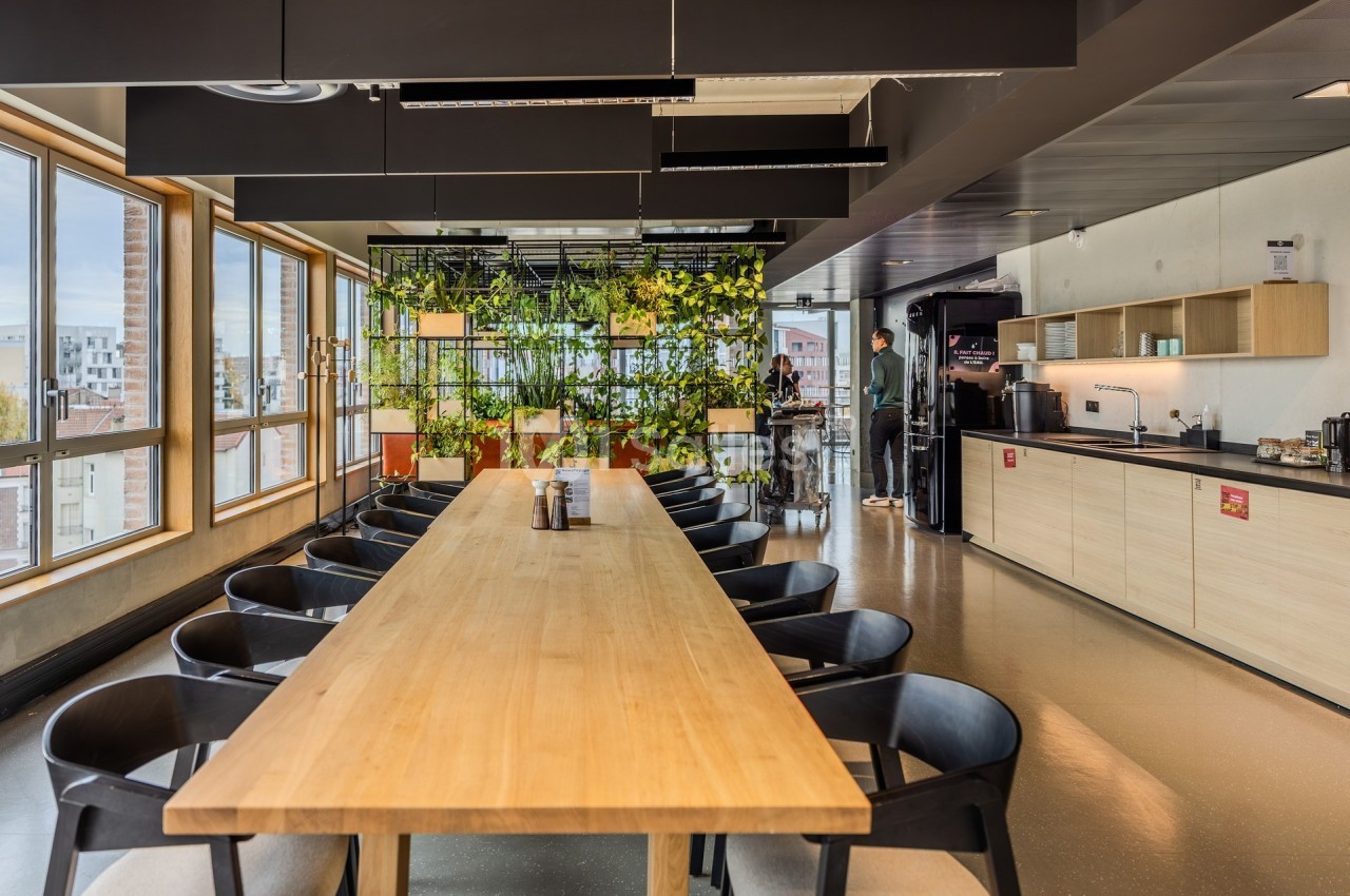 Salle de pause lumineuse avec une grande table en bois, des chaises noires, une cuisine équipée et des plantes suspendues.