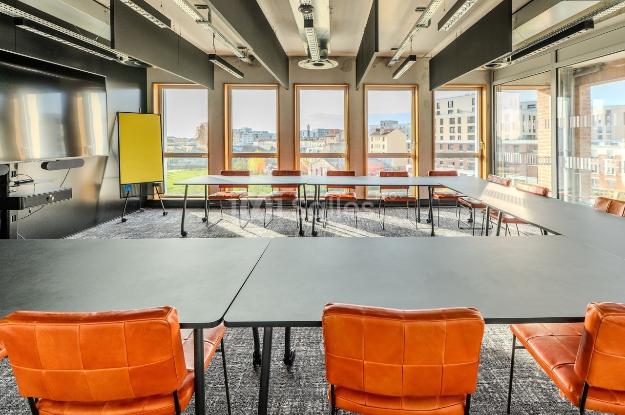 Salle de réunion lumineuse avec grandes fenêtres, tables noires, chaises orange et un tableau blanc.