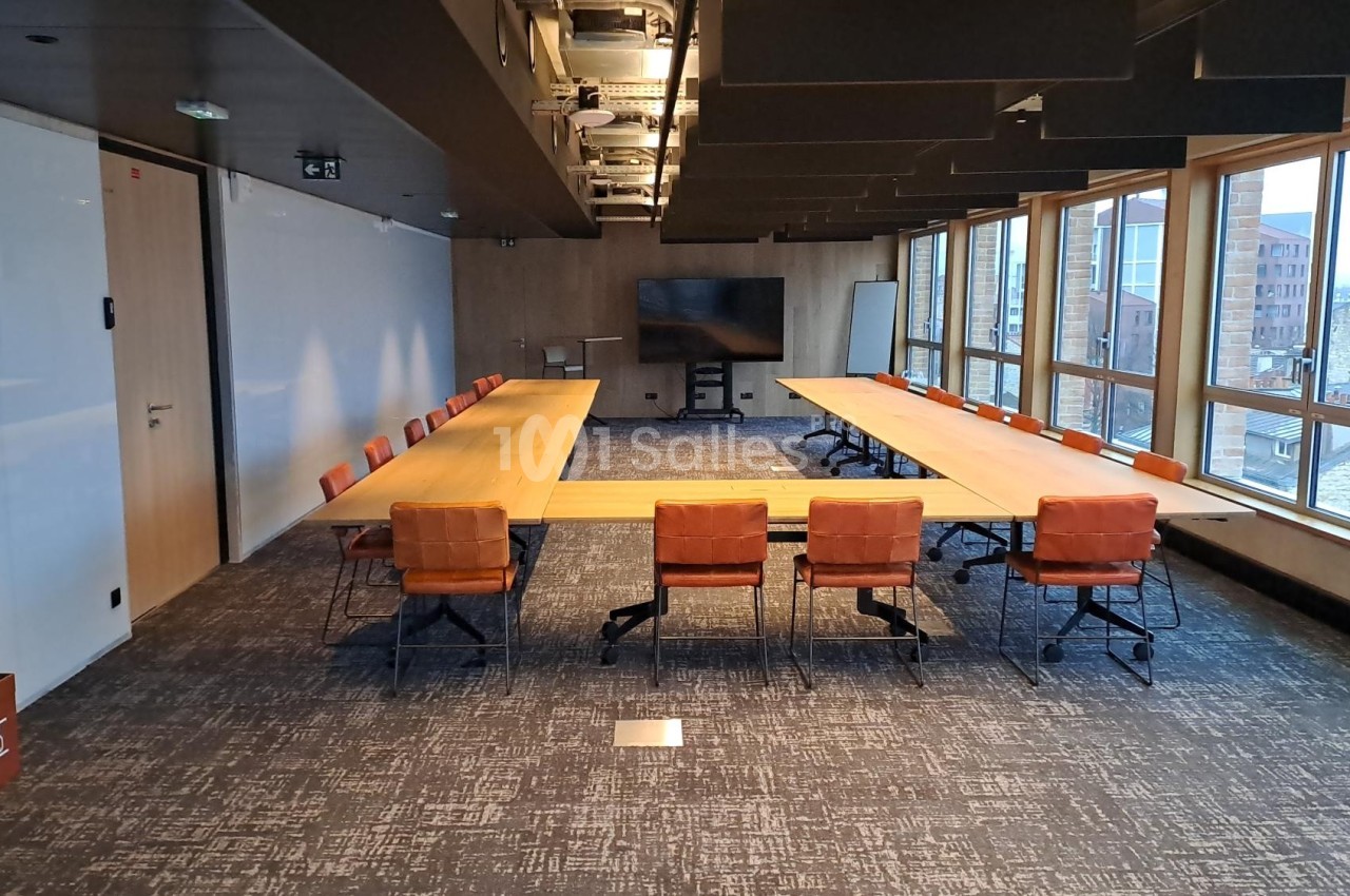 Salle de réunion moderne avec deux grandes tables en bois, chaises orange et baies vitrées donnant sur des bâtiments urbains.