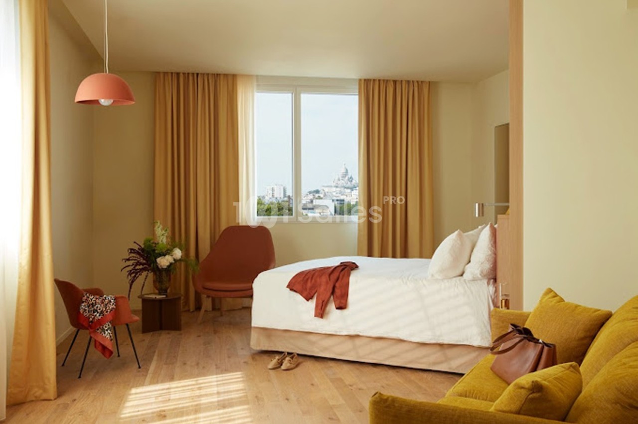 Chambre lumineuse avec lit double, fauteuils, rideaux beige, vue sur un bâtiment au loin par la fenêtre.