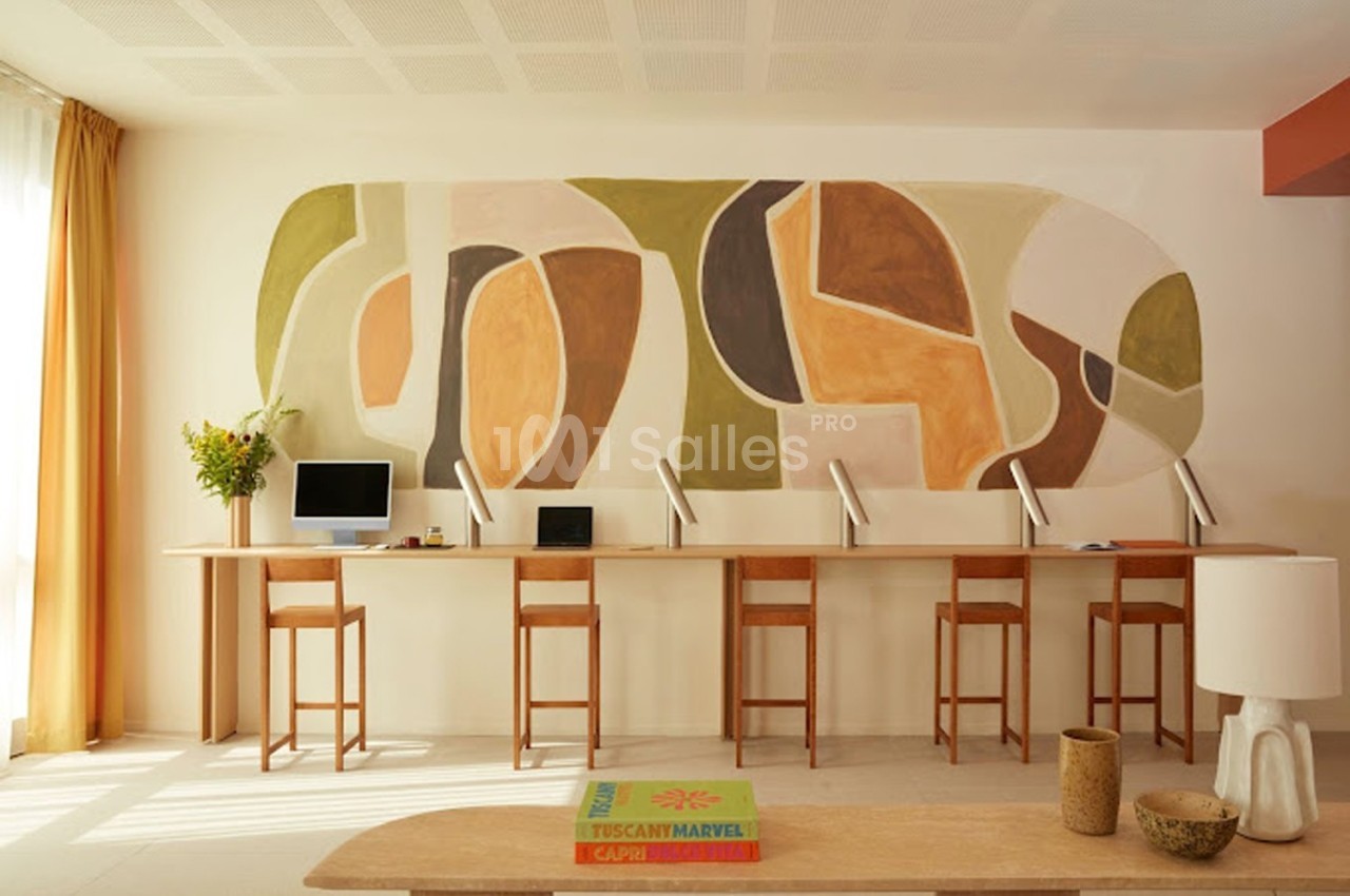 Espace de travail lumineux avec un long bureau, des chaises en bois et une fresque murale abstraite aux tons chauds.