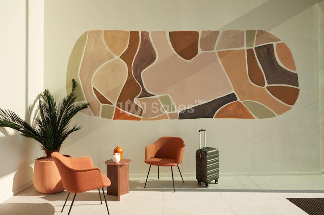 Mur décoré d'une fresque abstraite dans des tons terre, avec fauteuils orange, plante verte et valise posée au sol.