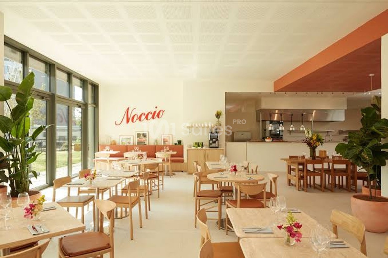 Salle lumineuse d'un restaurant moderne avec des tables en bois, des plantes et une cuisine ouverte en arrière-plan.