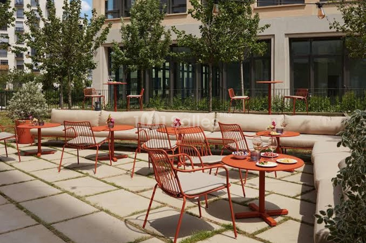 Terrasse extérieure avec tables et chaises rouges, entourée de verdure et bordée par un bâtiment moderne.