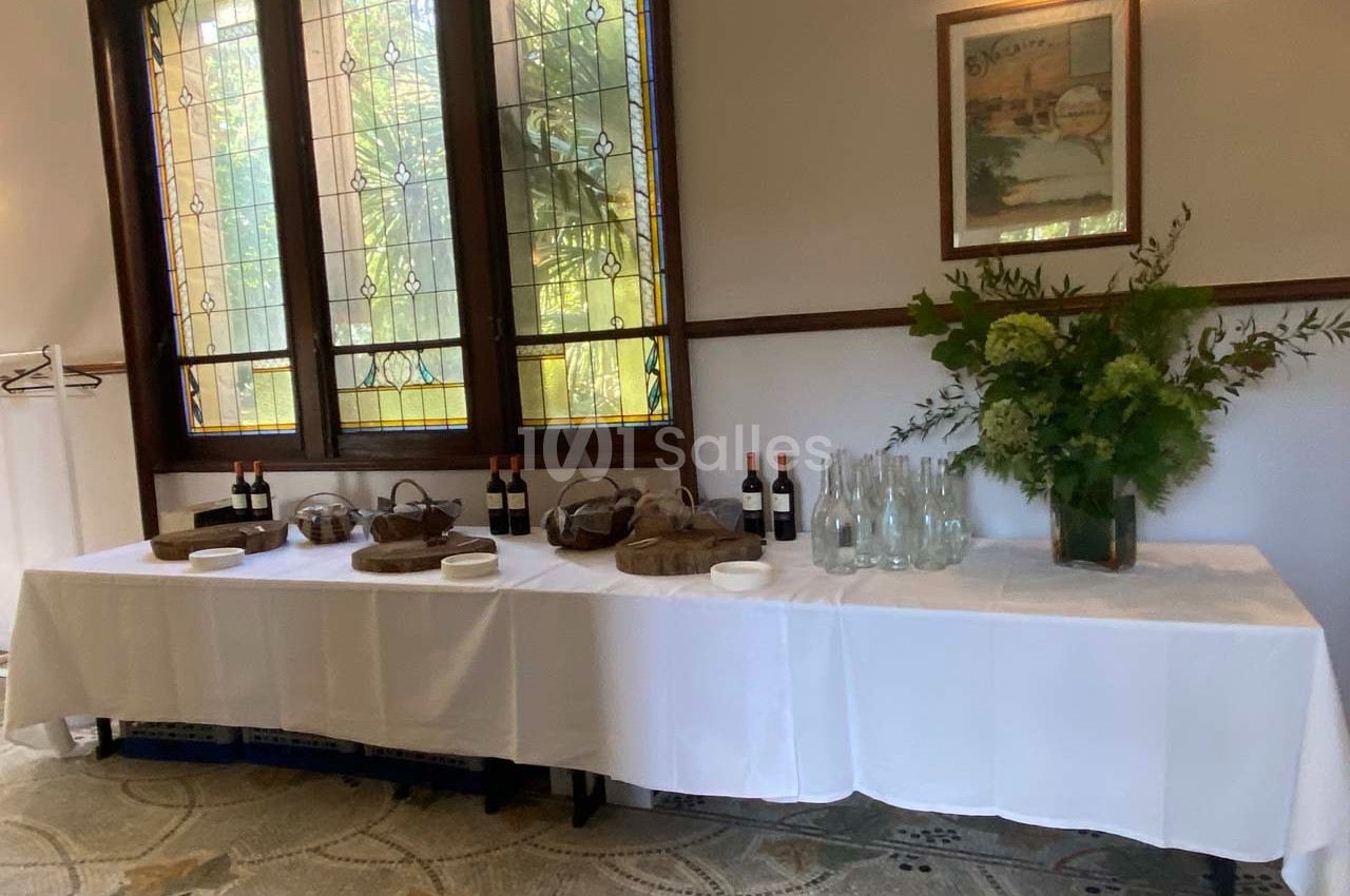 Table dressée avec nappes blanches, bouteilles de vin, carafes d'eau, paniers et décoration florale devant des vitraux.