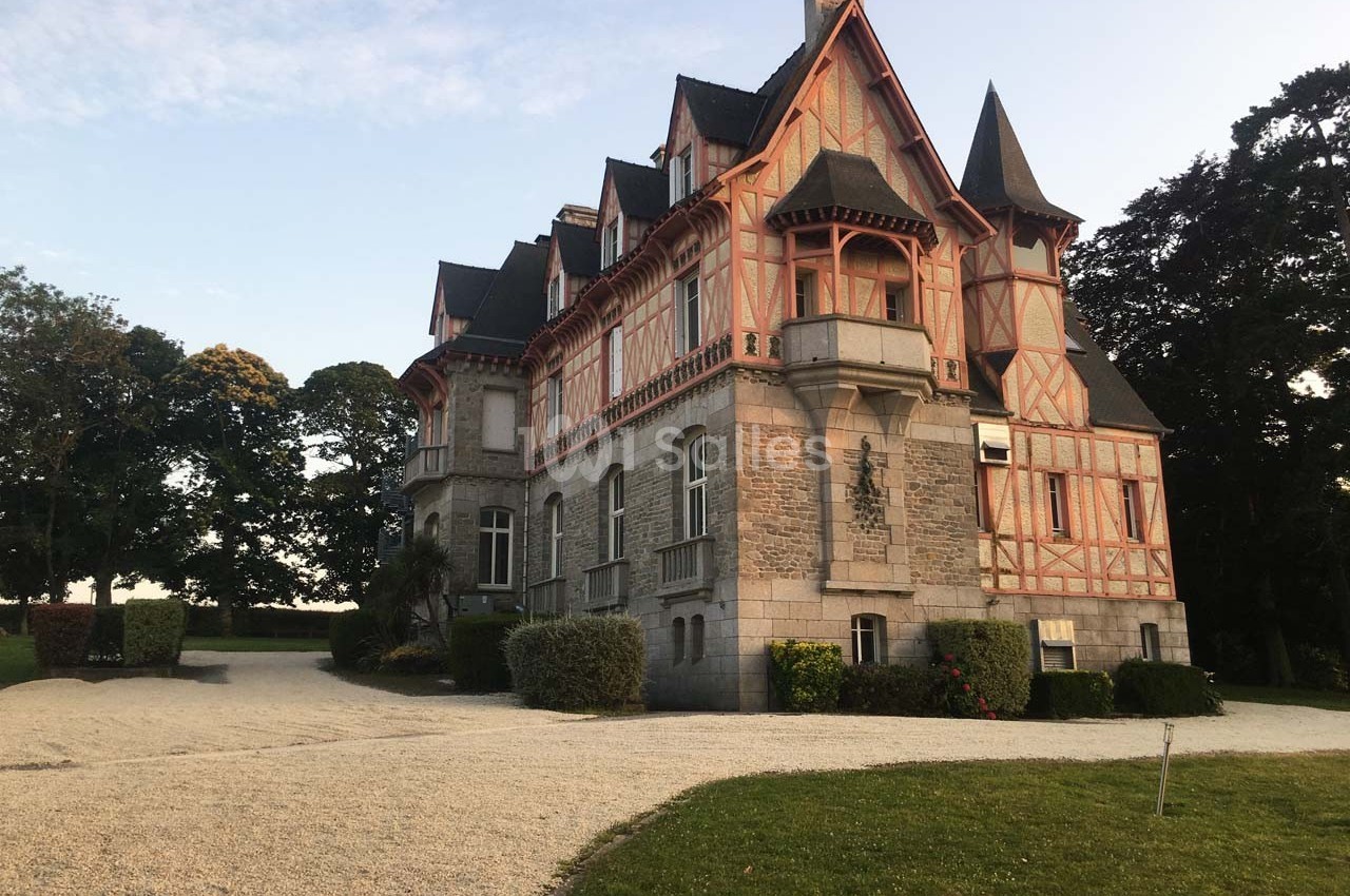 Grande maison en pierre et colombages, entourée de verdure, avec une allée gravillonnée au premier plan.