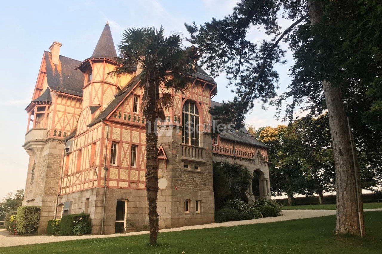Manoir en pierre et bois à colombages, entouré d'arbres, sous un ciel dégagé en fin de journée.