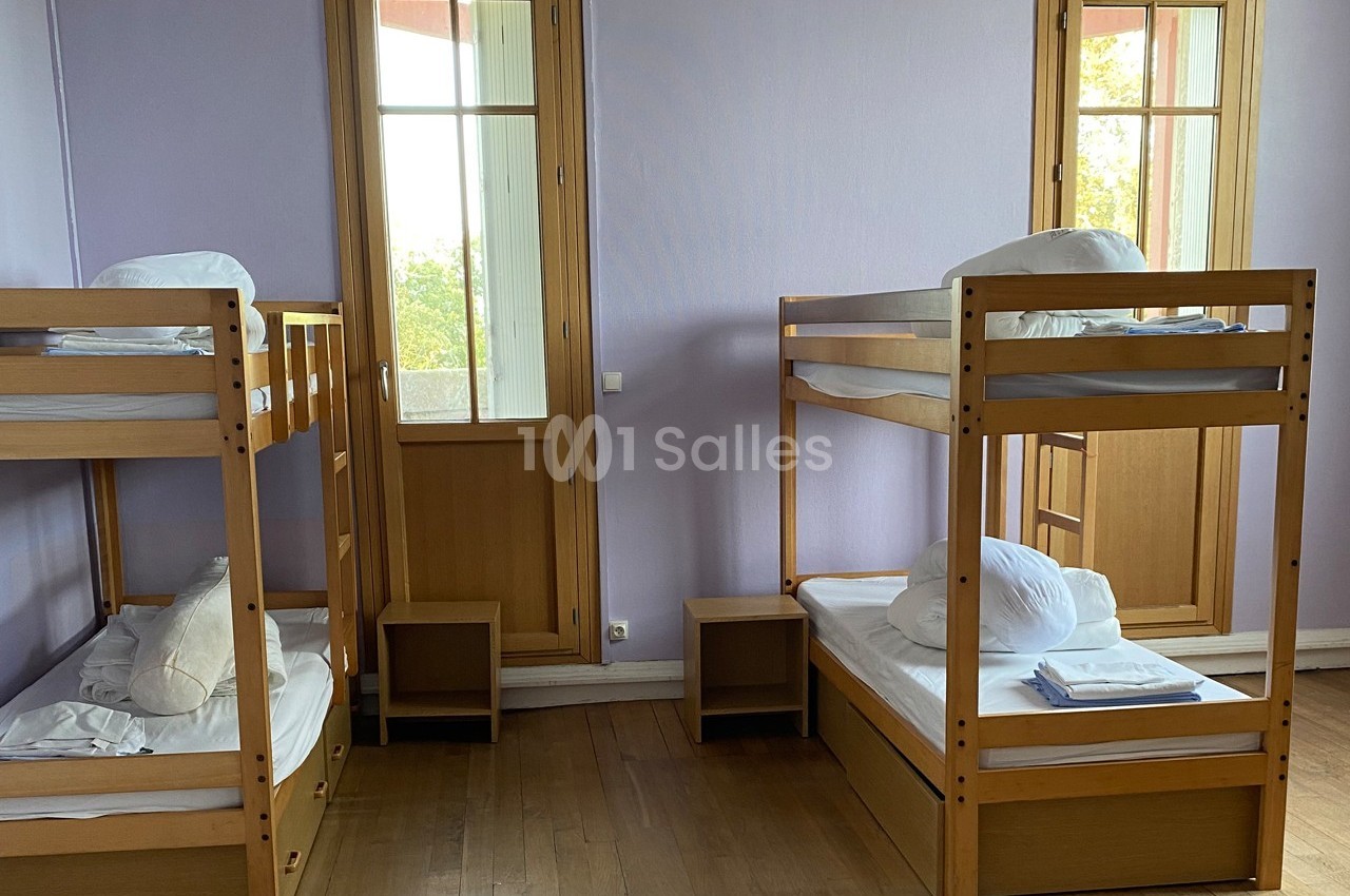 Chambre avec deux lits superposés en bois, draps blancs pliés, parquet au sol et fenêtres donnant sur l'extérieur.