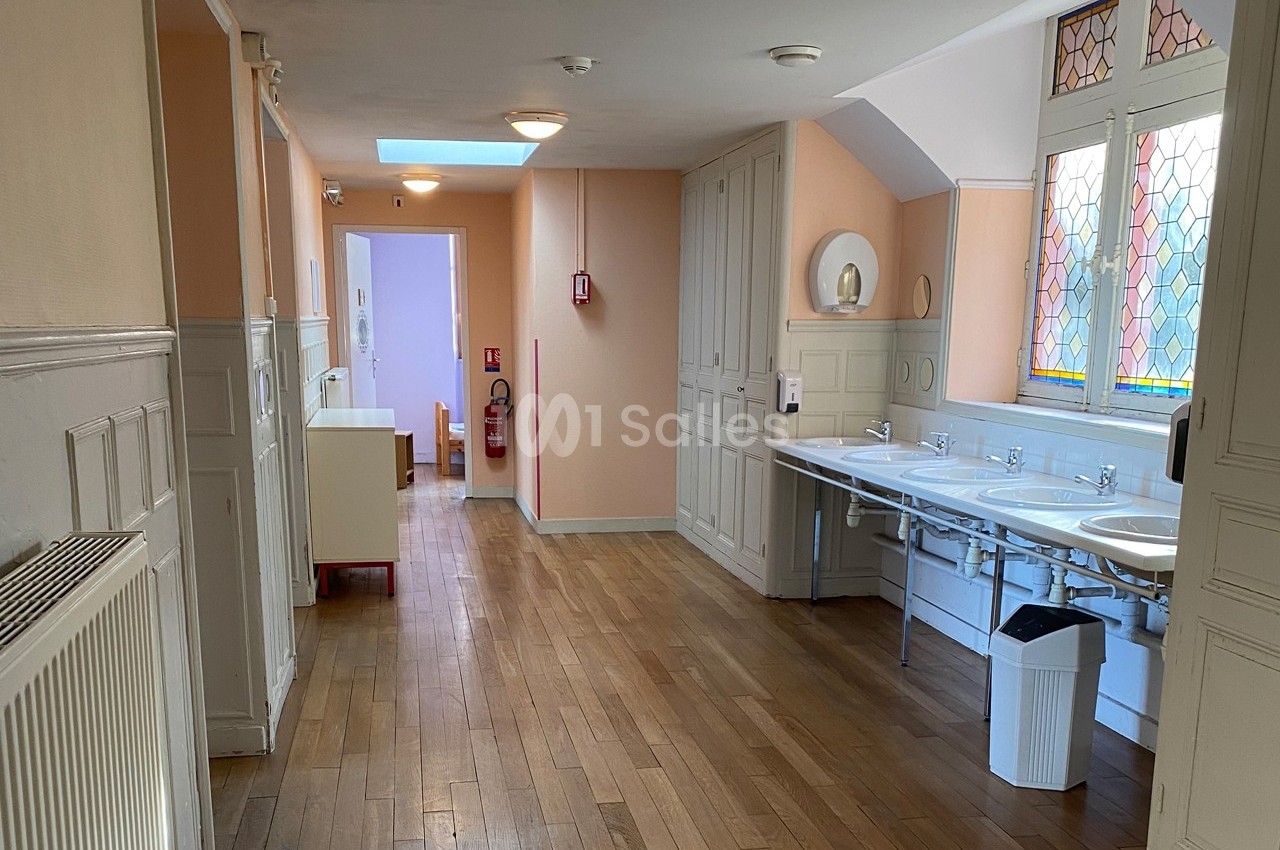 Couloir intérieur avec parquet, lavabos alignés à droite, vitraux colorés et mobilier clair en arrière-plan.