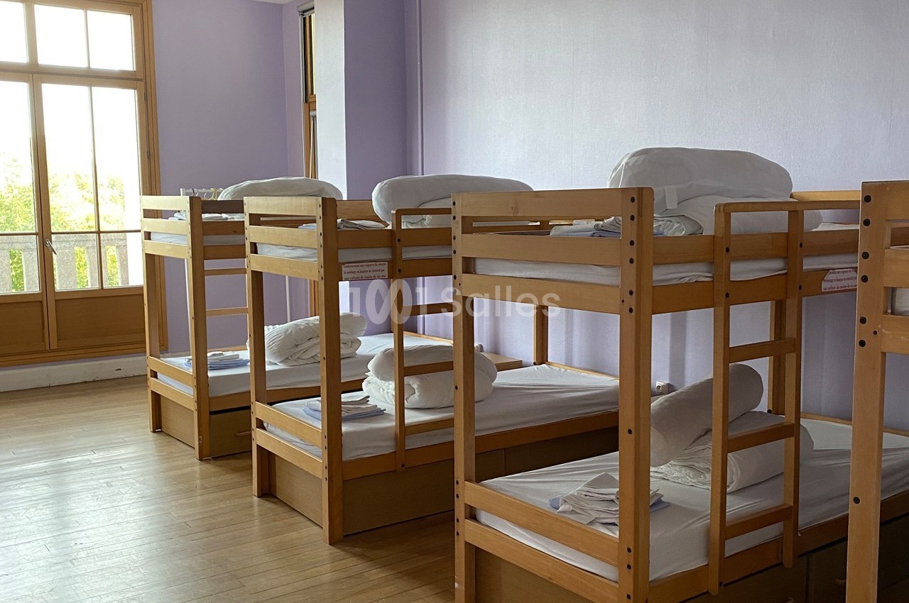 Dortoir avec plusieurs lits superposés en bois, draps blancs pliés, et grande fenêtre laissant entrer la lumière naturelle.