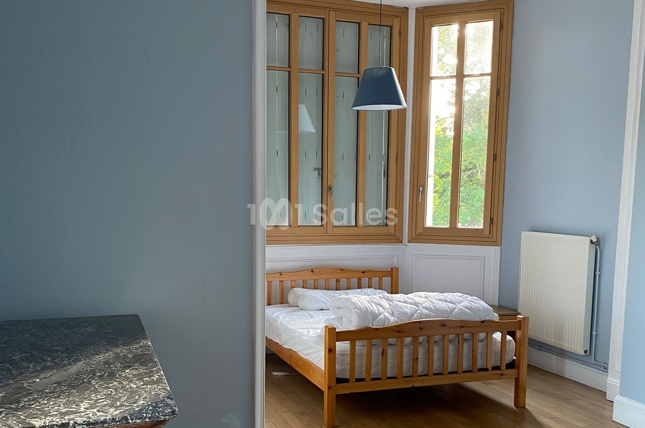 Chambre lumineuse avec un lit en bois, grande fenêtre à cadre en bois, murs bleus et parquet clair.