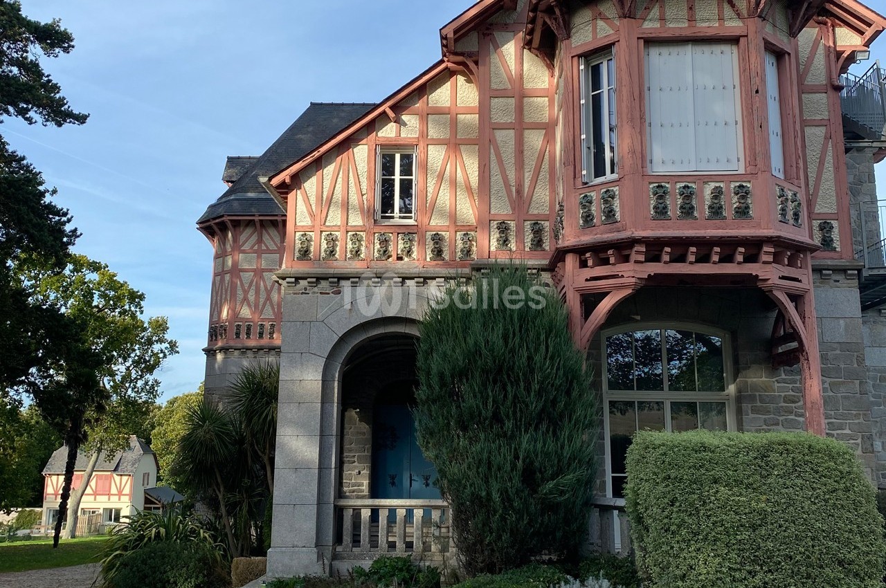 Façade d'une maison à colombages avec des détails en bois rouge, entourée de végétation et éclairée par la lumière du jour.