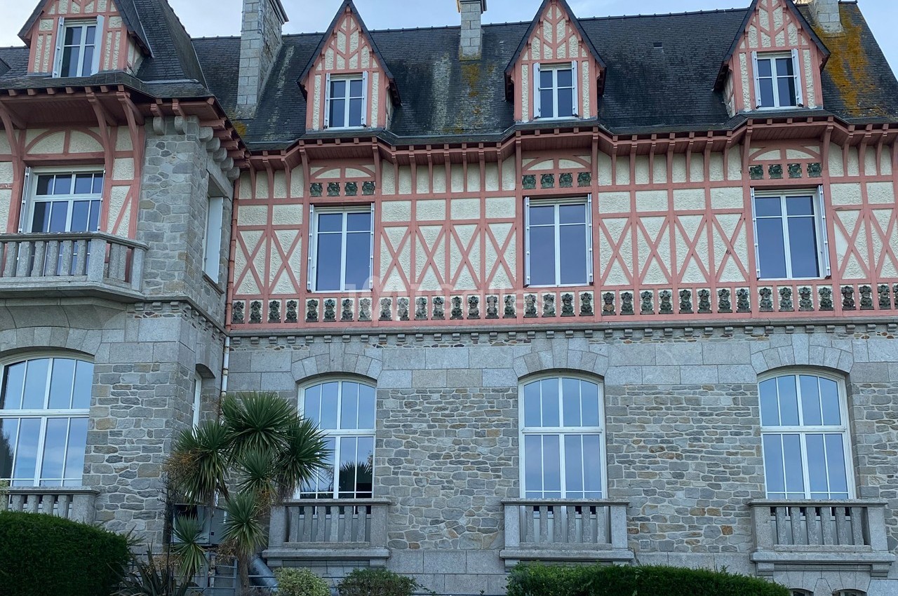 Façade d'une grande maison en pierre avec colombages roses, fenêtres à croisillons et toiture en ardoise.