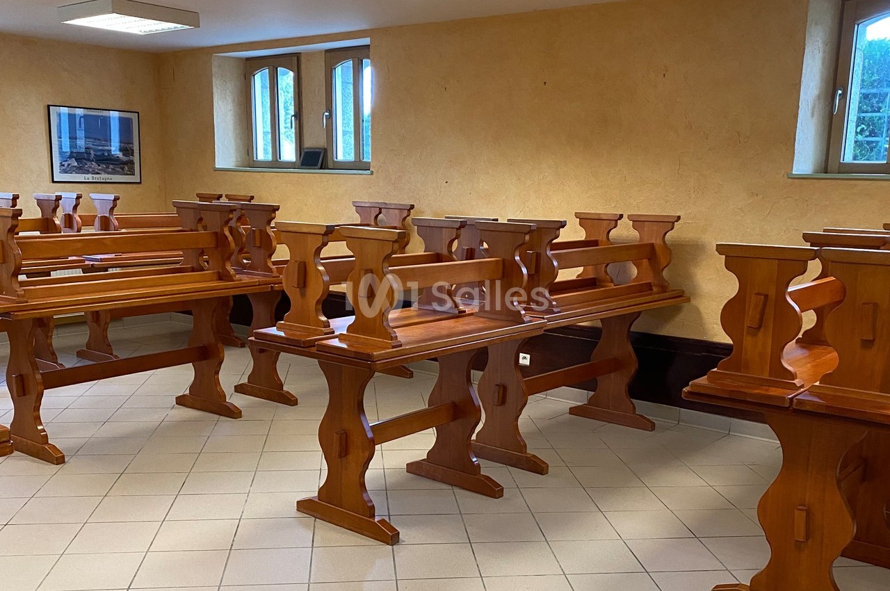 Salle avec des bancs en bois alignés sur un sol carrelé, éclairée par des fenêtres et des lampes au plafond.