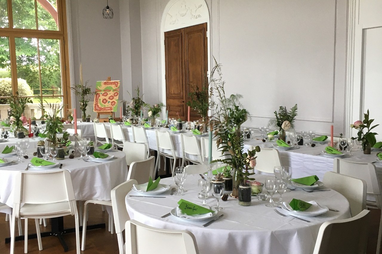Salle de réception décorée avec des tables rondes, nappes blanches, serviettes vertes et centres de table végétaux.