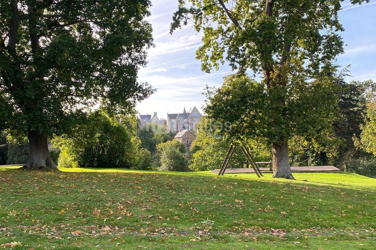 Pelouse parsemée de feuilles d'automne, entourée d'arbres, avec un bâtiment en pierre visible à l'arrière-plan.