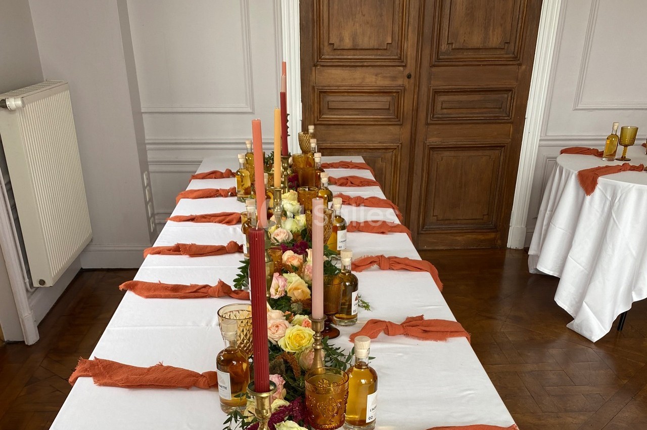 Table décorée avec des bougies colorées, des fleurs et des bouteilles ambrées, dans une pièce au parquet et porte en bois.
