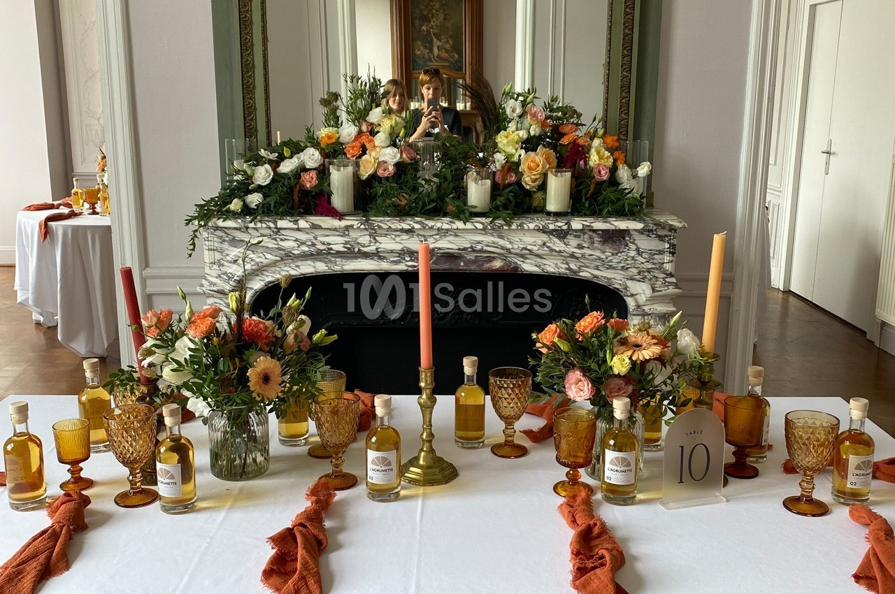 Table décorée avec des bougies, fleurs colorées et bouteilles, devant une cheminée ornée d'un miroir et d'arrangements…