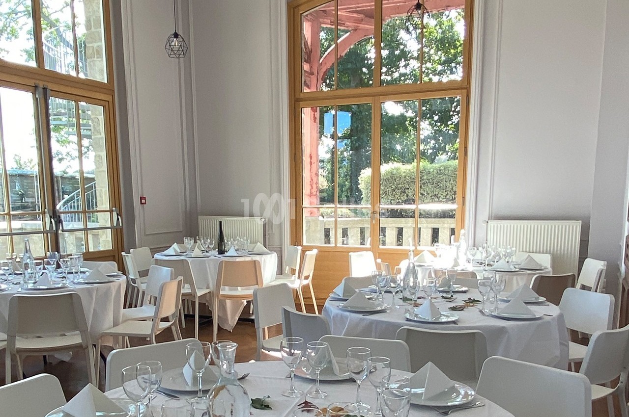 Salle lumineuse avec de grandes fenêtres, tables rondes dressées avec nappes blanches, verres et serviettes.