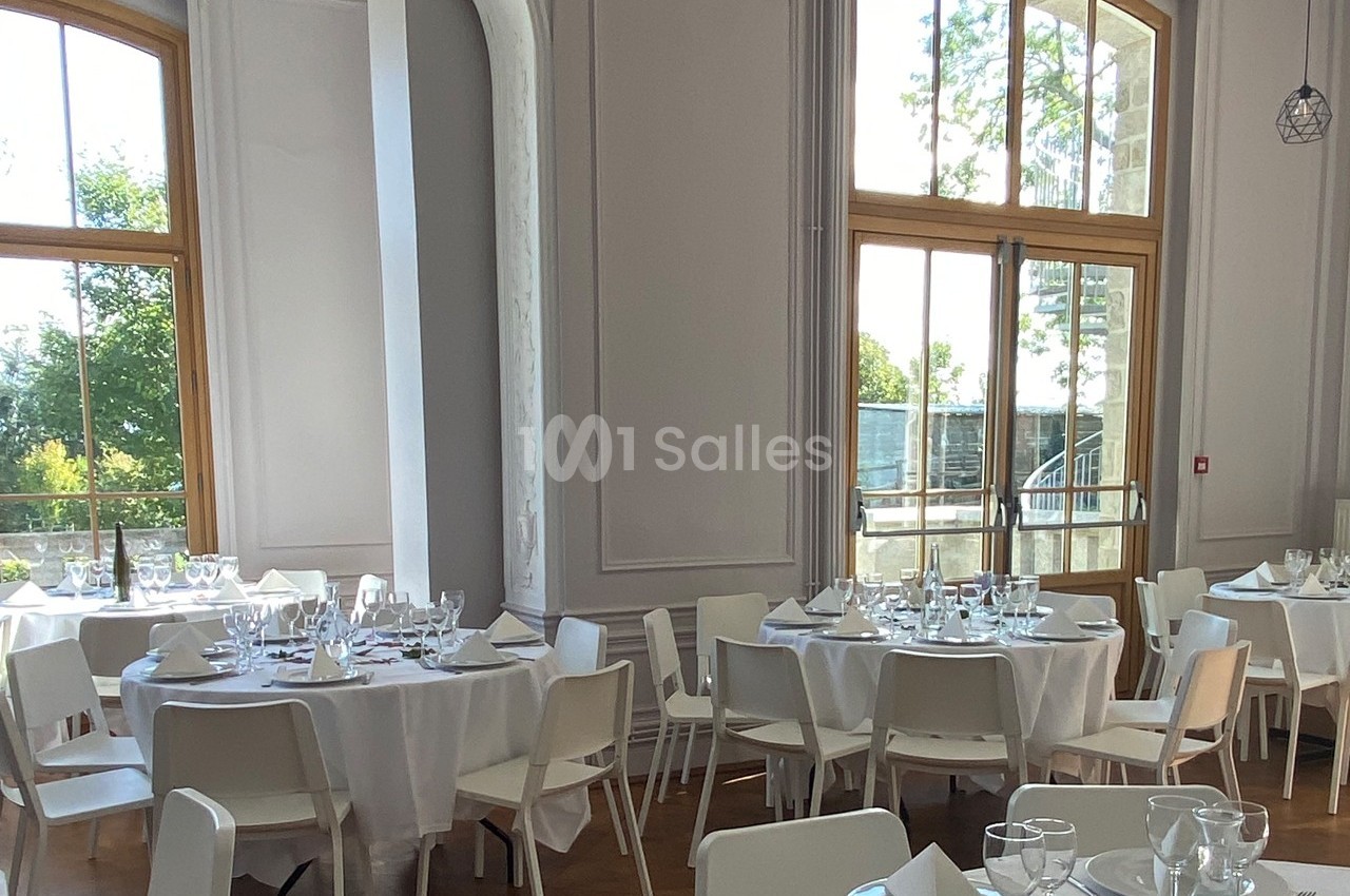 Salle lumineuse avec de grandes fenêtres, tables rondes dressées avec nappes blanches et chaises modernes.