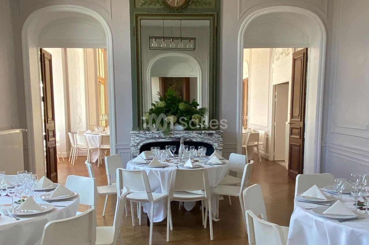 Salle élégante avec tables rondes dressées, chaises blanches et cheminée ornée de fleurs.