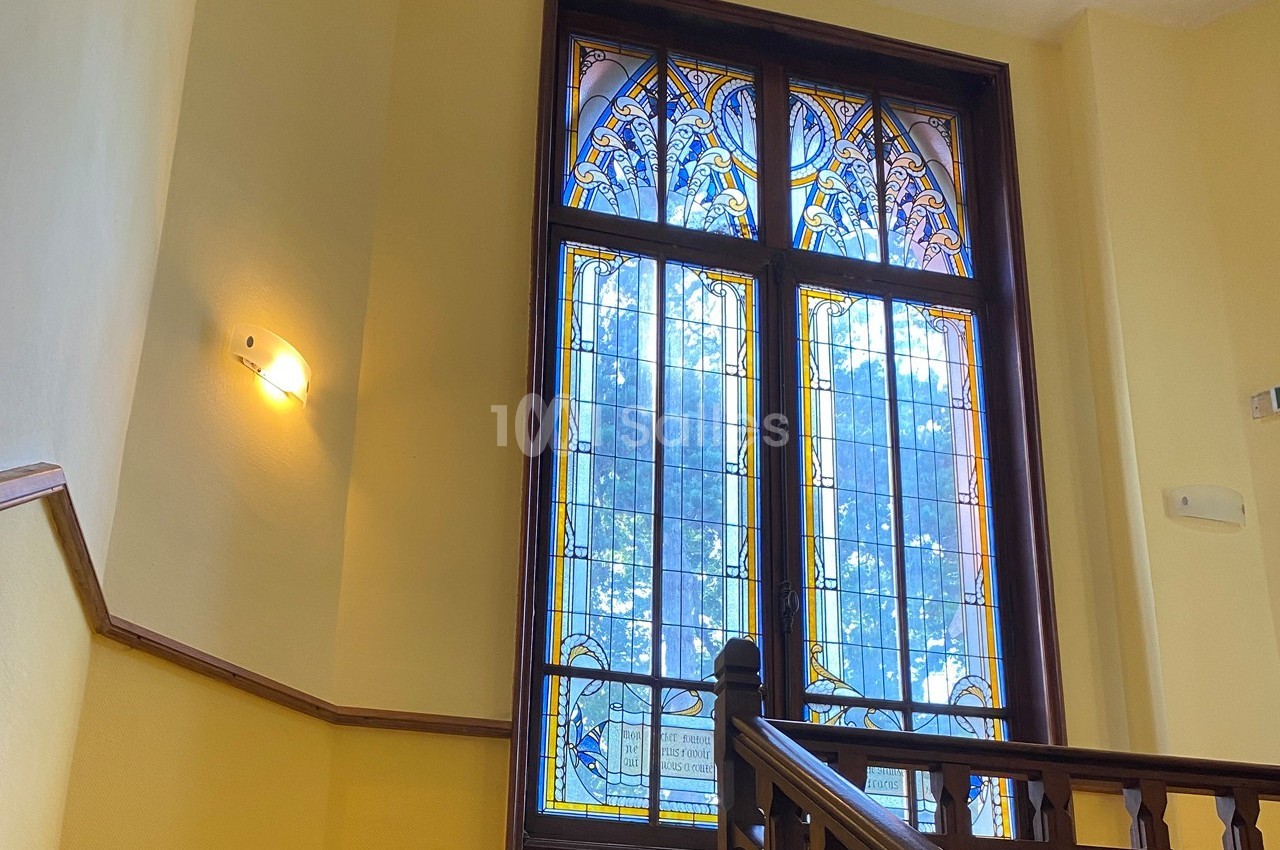 Fenêtre en vitrail coloré avec motifs décoratifs, encadrée de bois, éclairant un escalier intérieur.
