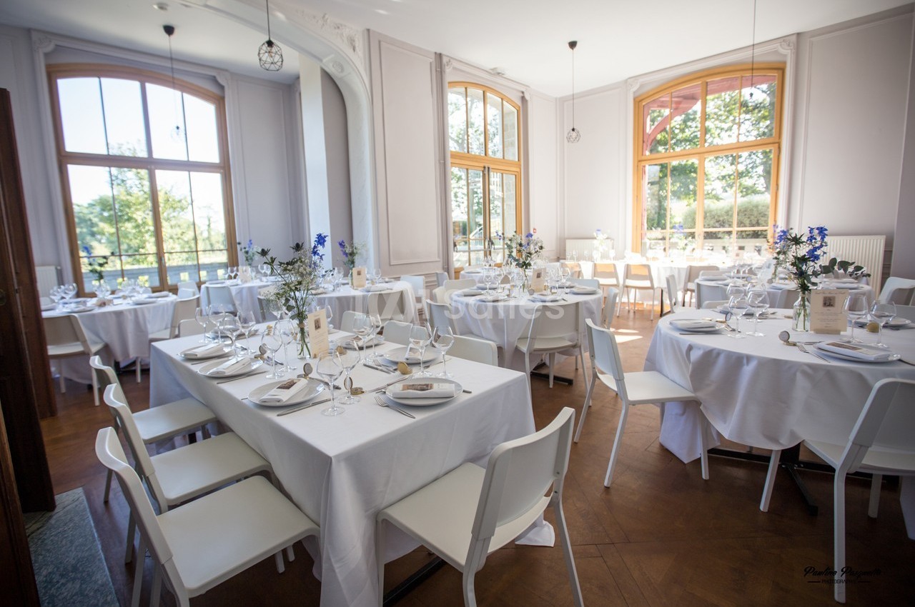 Salle lumineuse avec de grandes fenêtres, tables rondes et rectangulaires dressées pour un repas élégant.