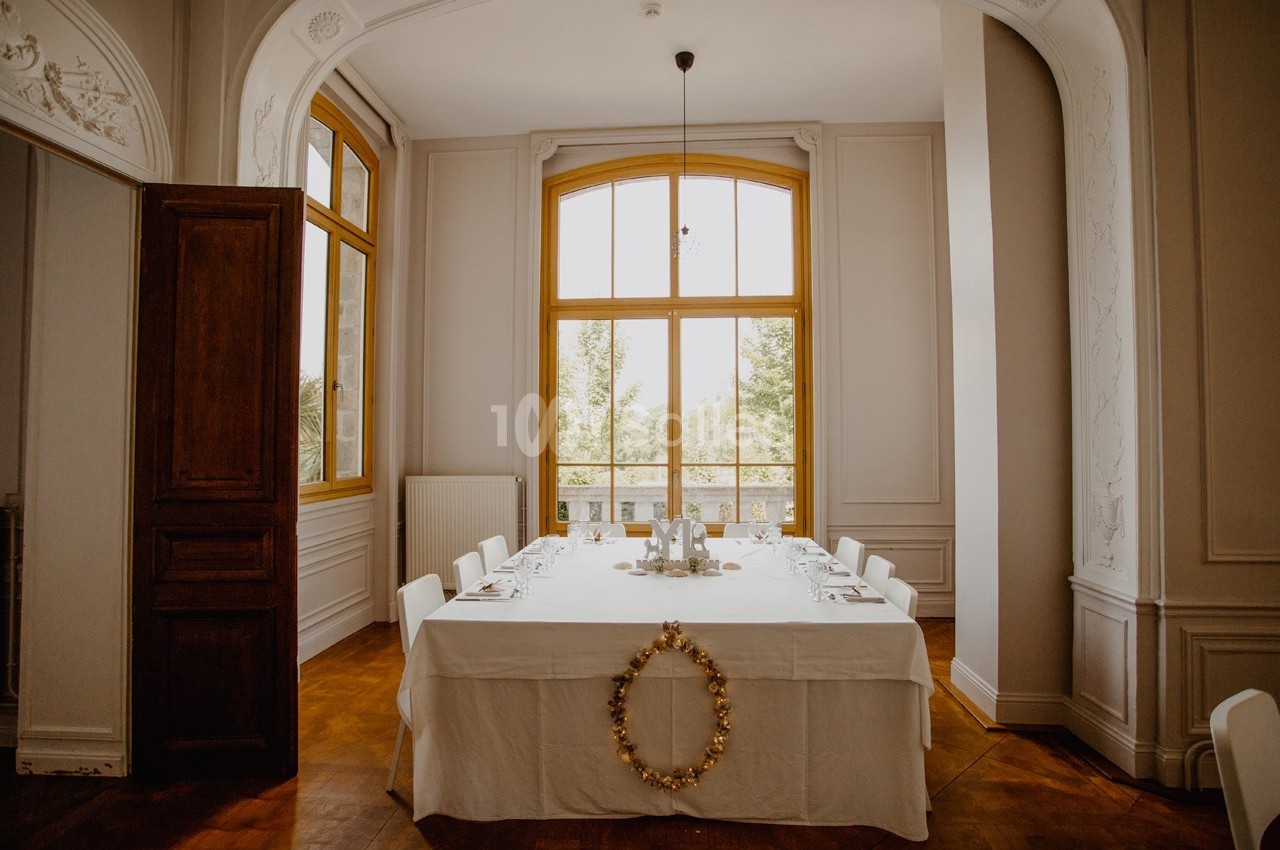 Salle à manger lumineuse avec une grande table dressée, entourée de chaises blanches, devant une fenêtre cintrée.
