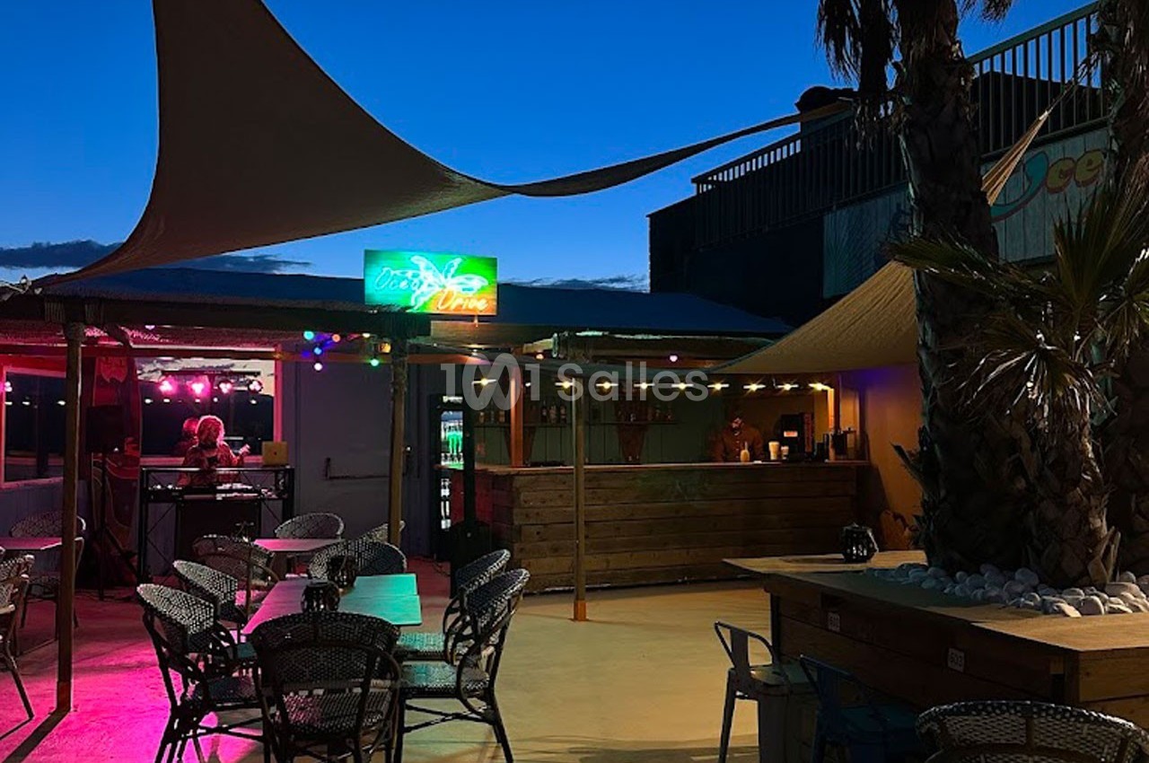 Terrasse extérieure éclairée en soirée avec des tables, des chaises, des voiles d'ombrage et un bar en arrière-plan.