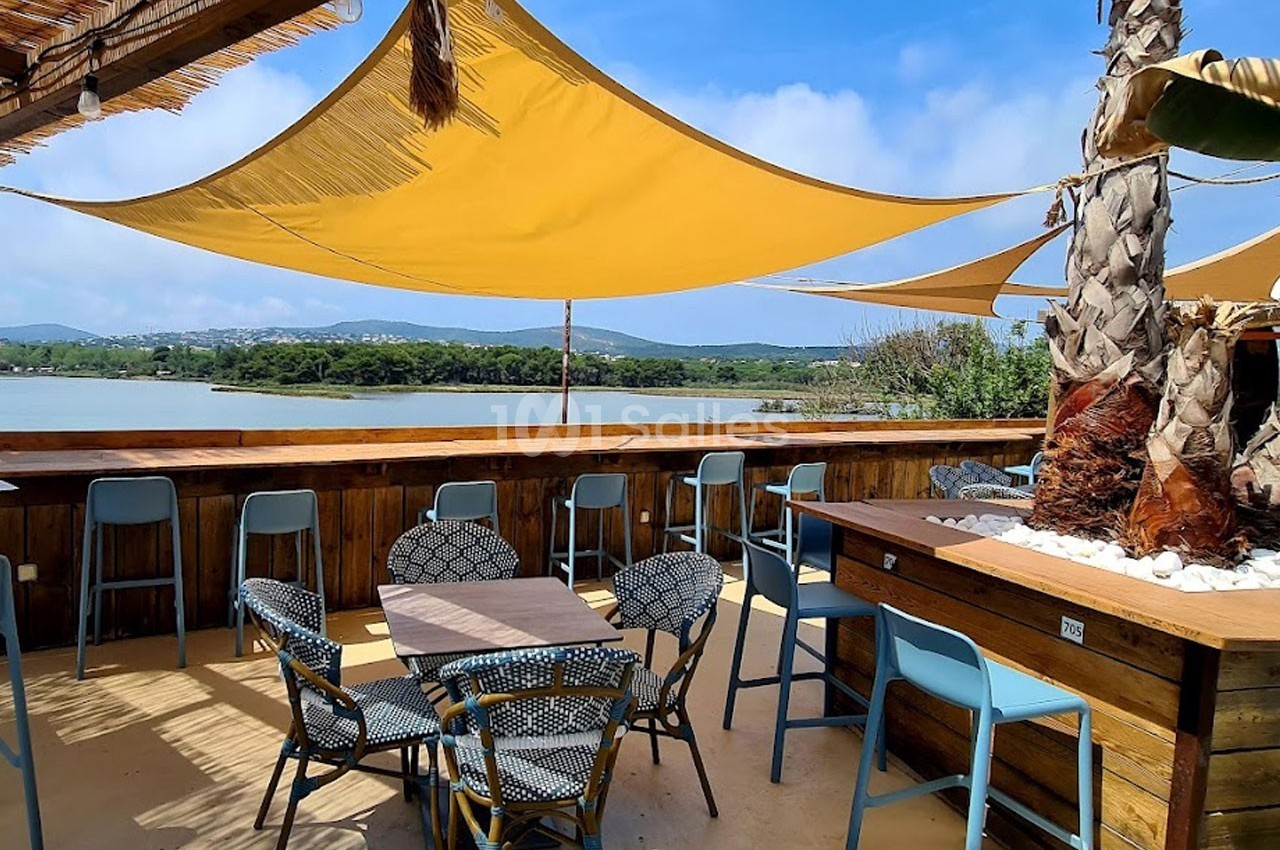 Terrasse en bois avec tables, chaises et voiles d'ombrage, offrant une vue sur un lac et des collines verdoyantes.