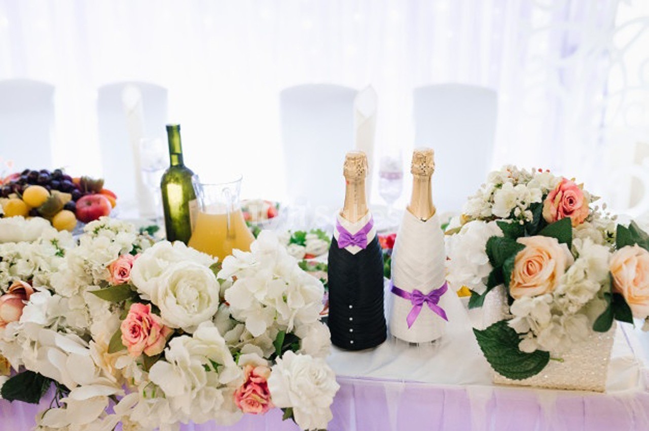 Table décorée avec des fleurs blanches et roses, bouteilles habillées, fruits et boissons disposés pour une réception.