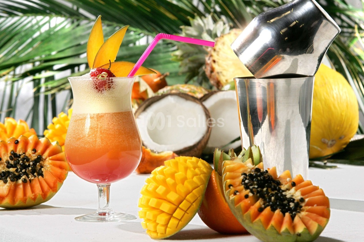 Cocktail exotique avec mousse, décoré de fruits tropicaux, à côté d'un shaker et de fruits frais sur une table blanche.