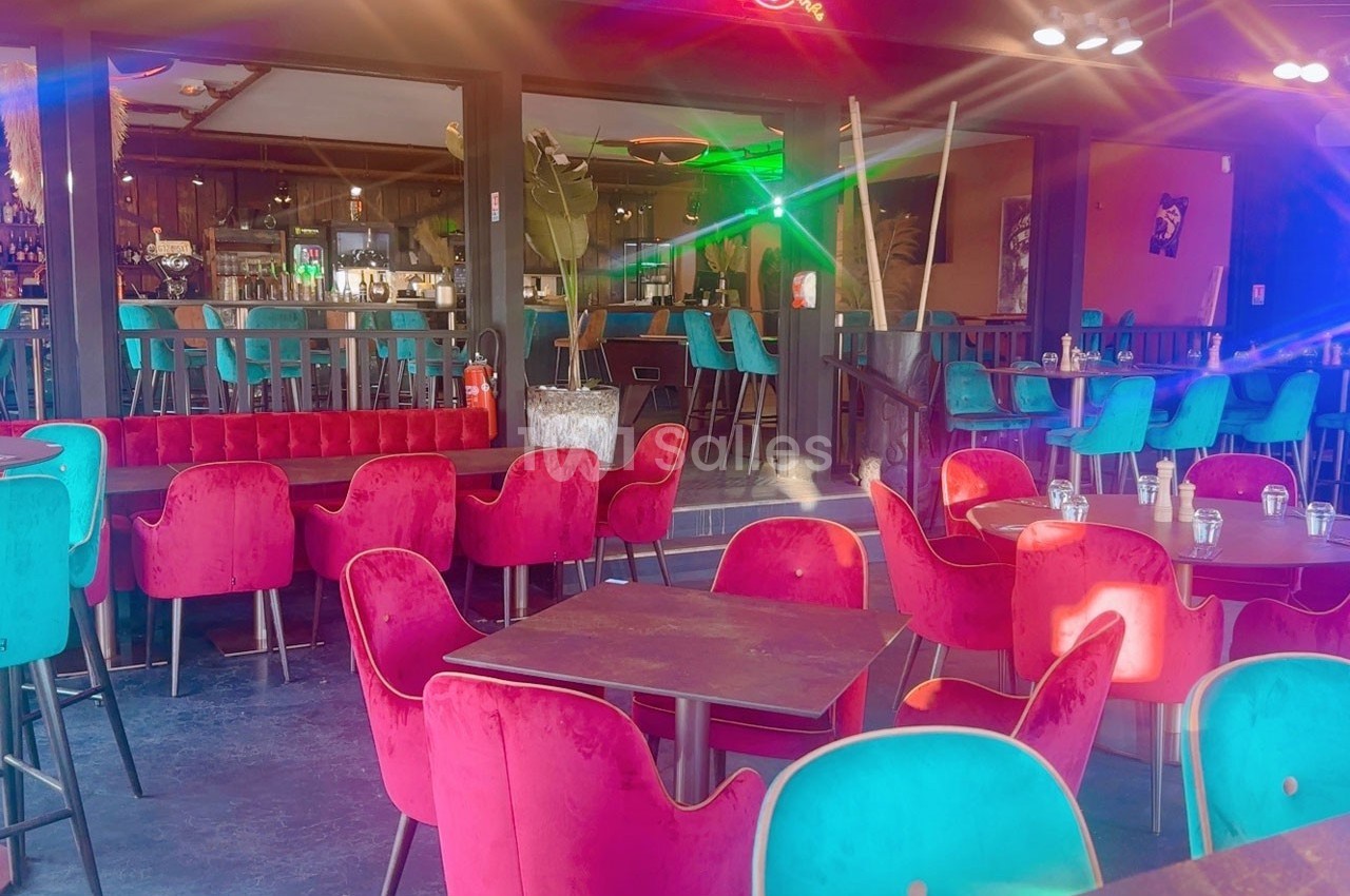 Salle de restaurant colorée avec des chaises rouges et turquoise, tables en bois et un bar en arrière-plan.