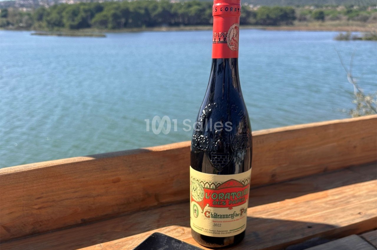 Bouteille de vin rouge posée sur une table en bois, avec un lac et des collines en arrière-plan.