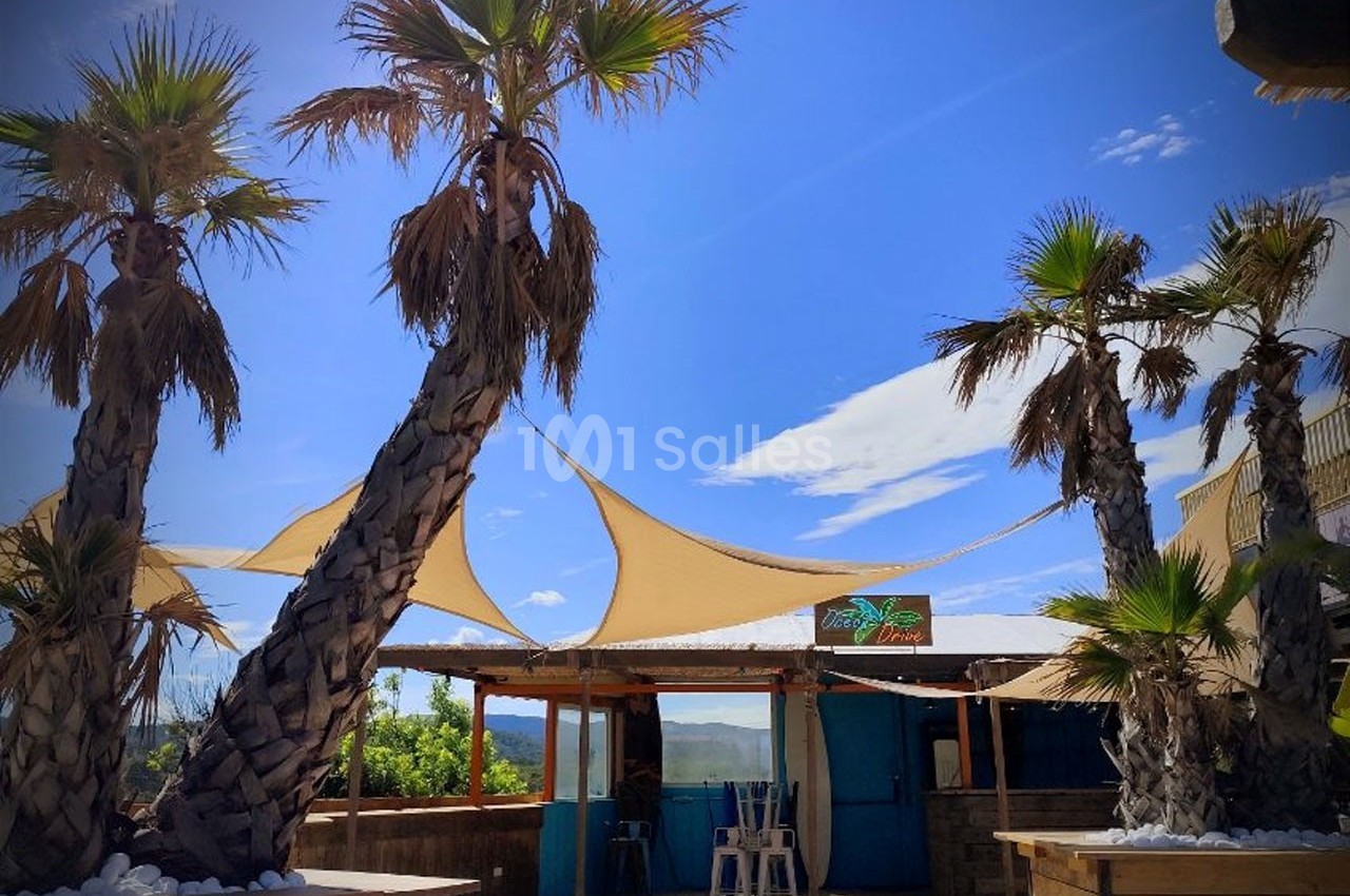 Terrasse extérieure avec palmiers, voiles d'ombrage beige et cabane en bois sous un ciel bleu.